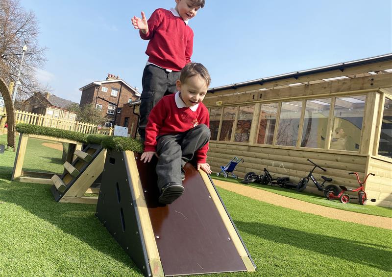 Two children are standing on a ramped wooden block from the Get Set, Go! Snowdonia set. The child at the front is sliding down the ramp whilst the child behind is stood up and watching the other child.
